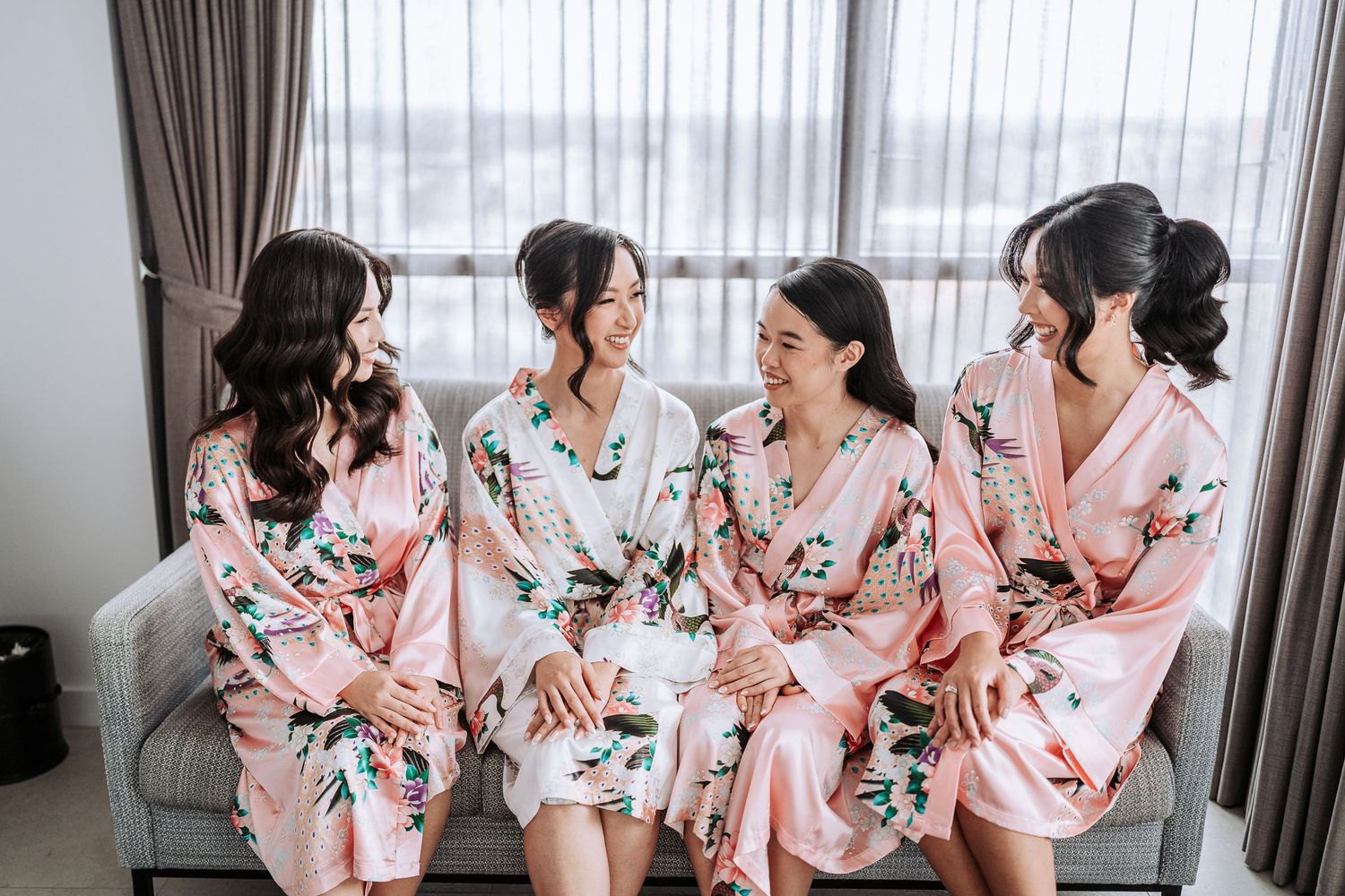 Bridesmaids wearing matching floral pink robes laugh together while getting ready for a wedding celebration.
