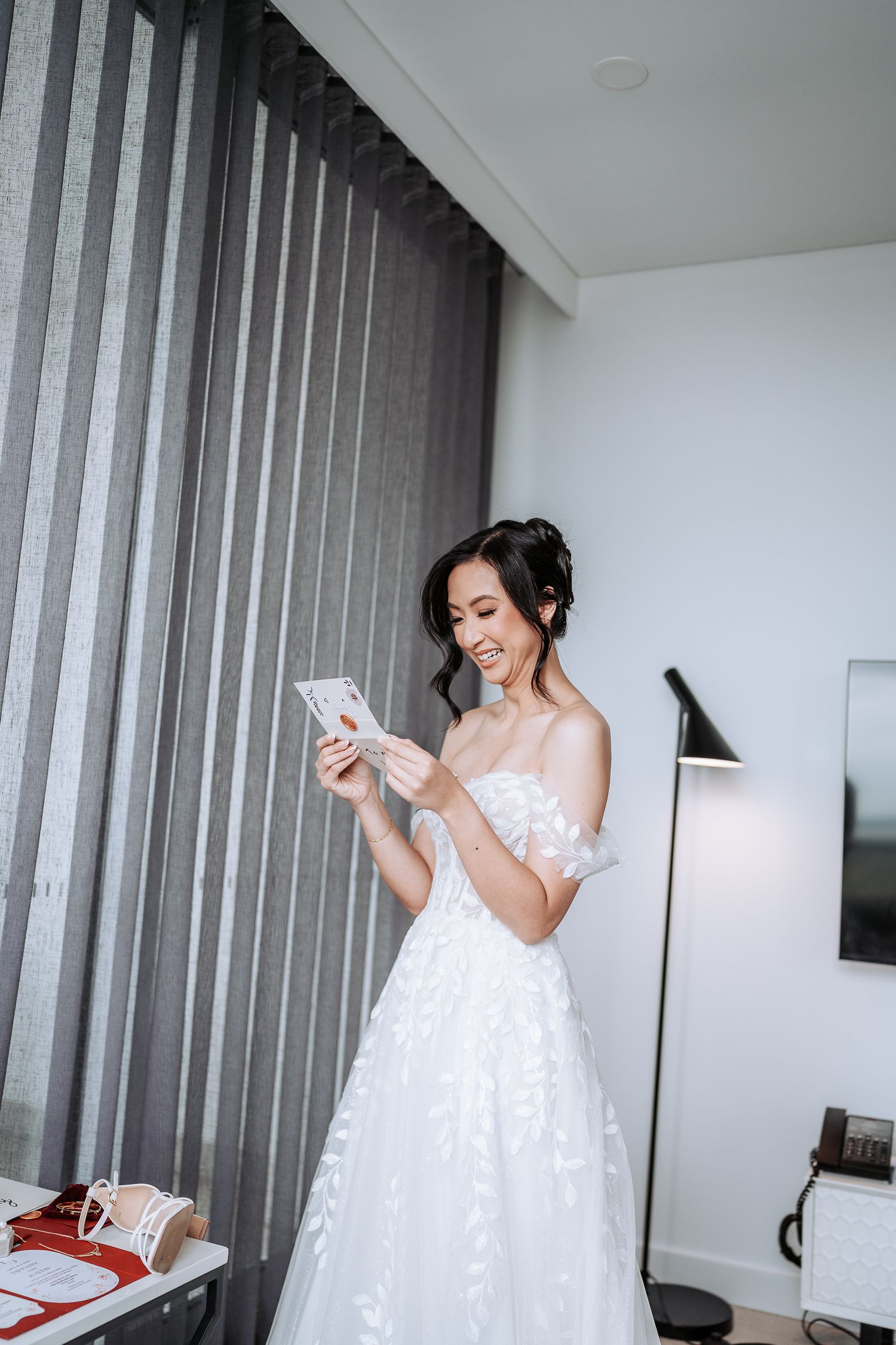 A bride in a white lace dress getting ready in a modern hotel room with vertical blinds.
