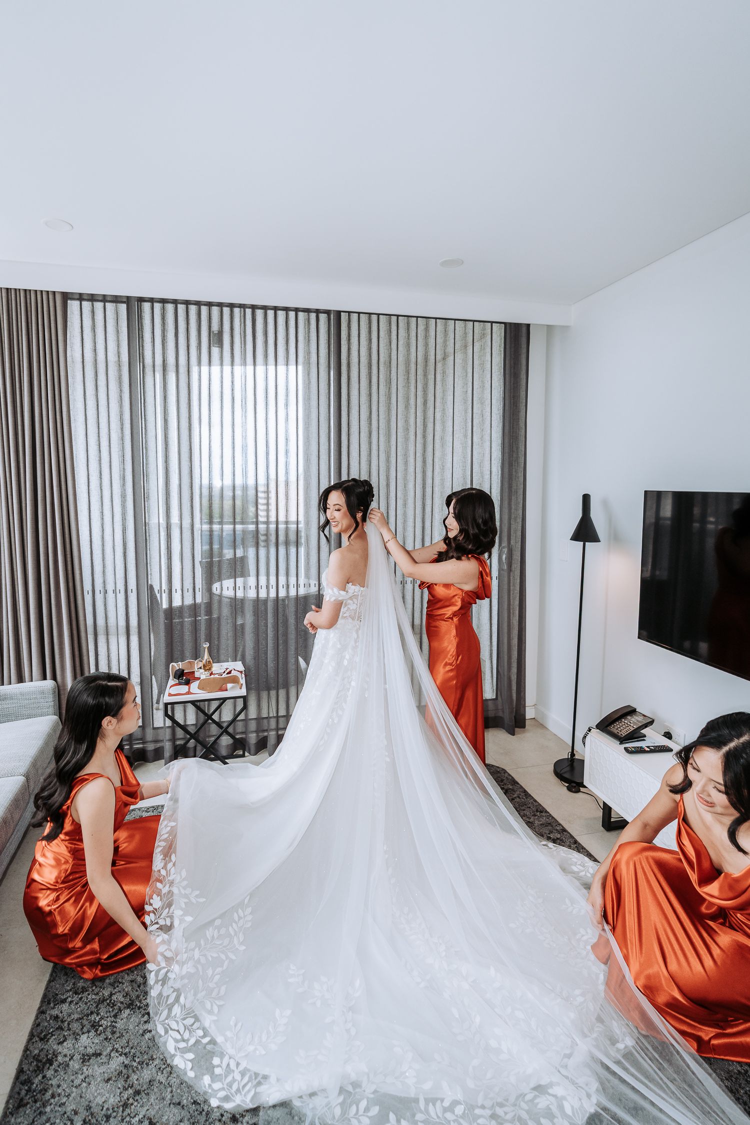 Bridesmaids in coral dresses admire a flowing white wedding gown with lace details in a modern hotel room.