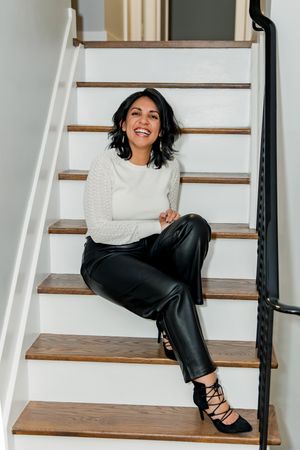 Person in white sweater and black pants poses on wooden stairs in bright interior setting.