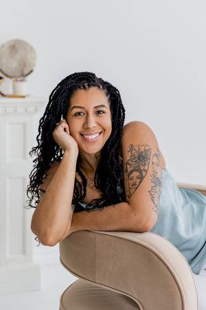 A person with long braids and a floral arm tattoo relaxes on a beige chair while smiling at the camera.