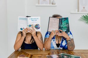 Four people hold up different books in front of their faces while sitting at a wooden table.