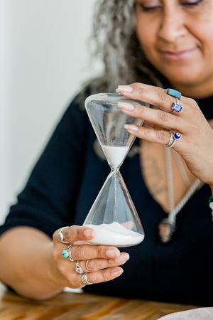 A person wearing black holds a small hourglass timer while sitting at a wooden table.