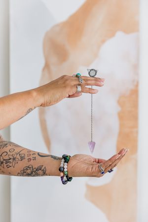 Tattooed hands holding a crystal pendulum with multiple rings and bracelets against a light background.