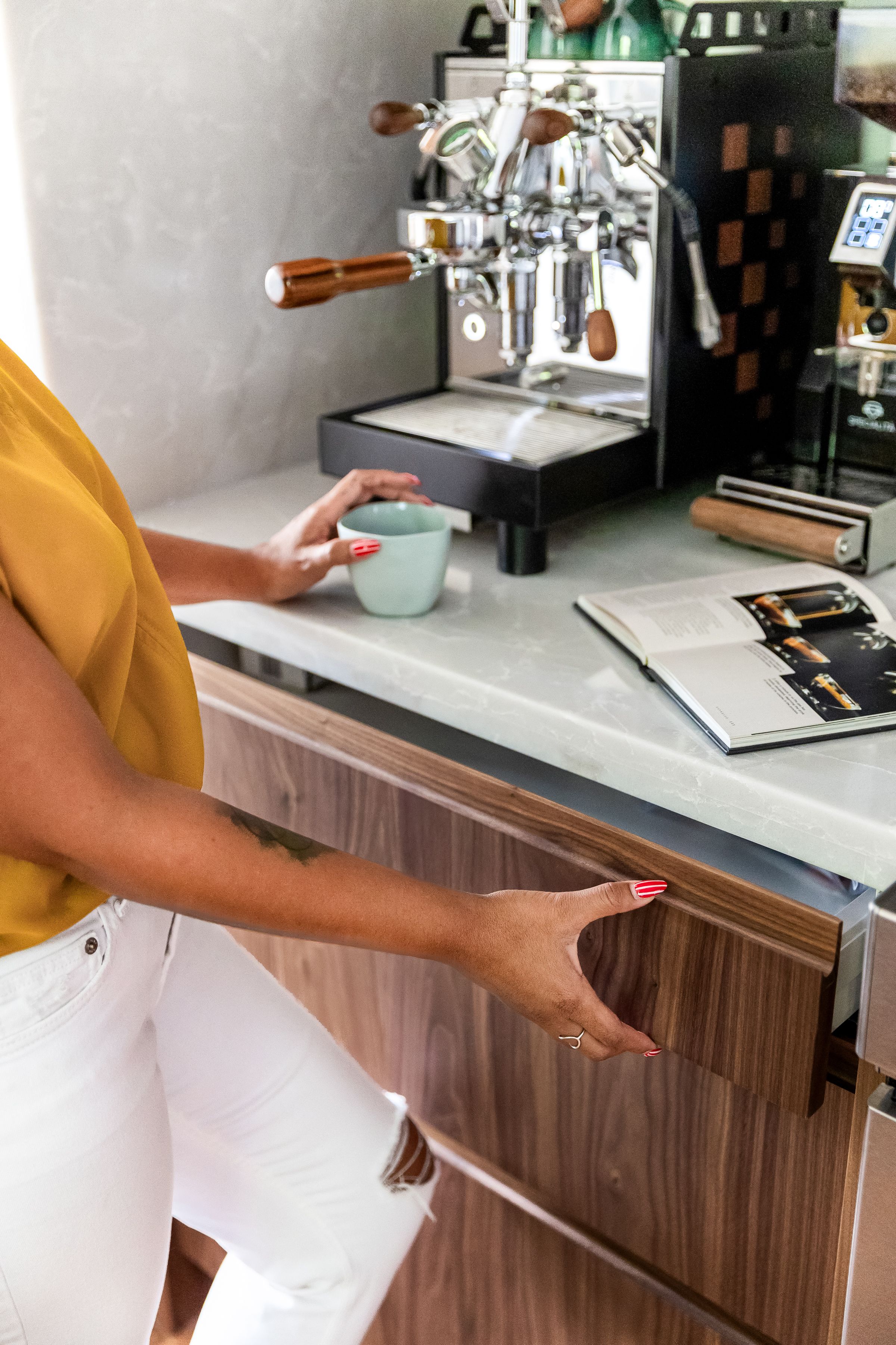 Hand opening a modern wooden cabinet drawer in a sleek kitchen with white countertops and coffee equipment.