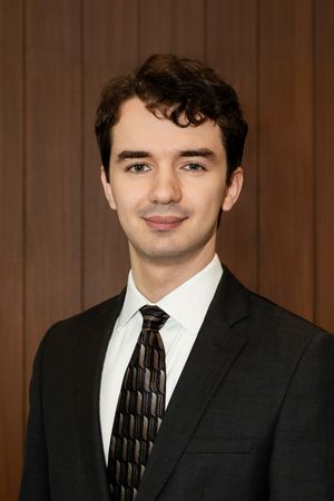 A person wearing a black suit and patterned tie stands against a wood paneled wall in a formal portrait setting.