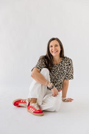 Person in leopard print shirt and white pants relaxes on white floor with red slippers in casual photo shoot.