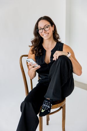 Professional woman in black outfit and glasses sits casually on wooden chair while checking phone and smiling.