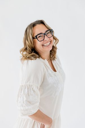 Person wearing glasses and a white blouse smiles warmly against a light background in a studio portrait series.