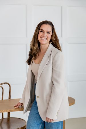 Professional wearing beige blazer and jeans poses against white wall in casual business attire.