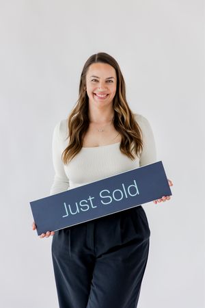 A person in a white sweater holds up a navy blue sign while smiling against a light background.