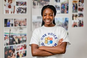 A person wearing a white One Voice t-shirt stands smiling with arms crossed in front of a photo wall.