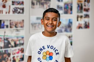 Smiling young boy wearing a white One Voice t-shirt standing in front of a wall of photos.