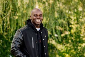 A person wearing a black jacket smiles while standing outdoors in a natural green setting.