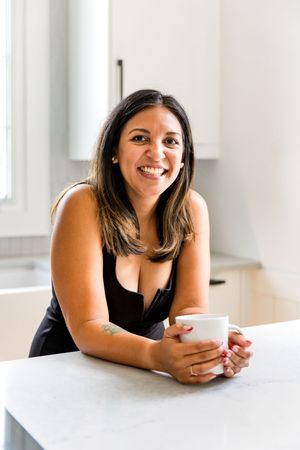 Woman in black tank top smiling while relaxing at white counter with coffee cup.