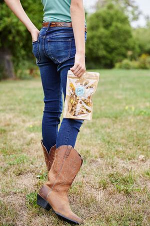 Person wearing brown cowboy boots and blue jeans holding a snack bag while standing in a grassy field.