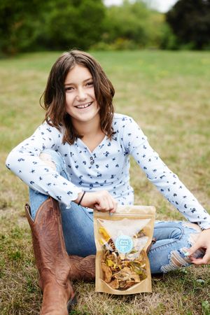 Someone wearing a white patterned shirt and cowboy boots sits in grass while displaying a bag of snacks.