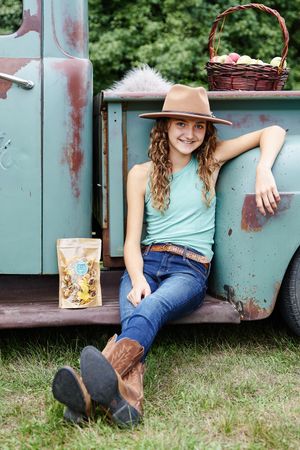 Rustic country style portrait with vintage truck, brown cowboy boots, jeans and teal tank top against green foliage.