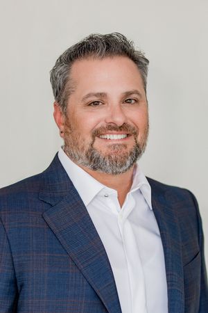 Professional headshot of someone wearing a navy plaid blazer and white dress shirt against a light gray background.