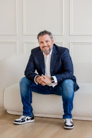 A person in a navy blazer and jeans sits casually on a light-colored couch in a bright, minimalist setting.