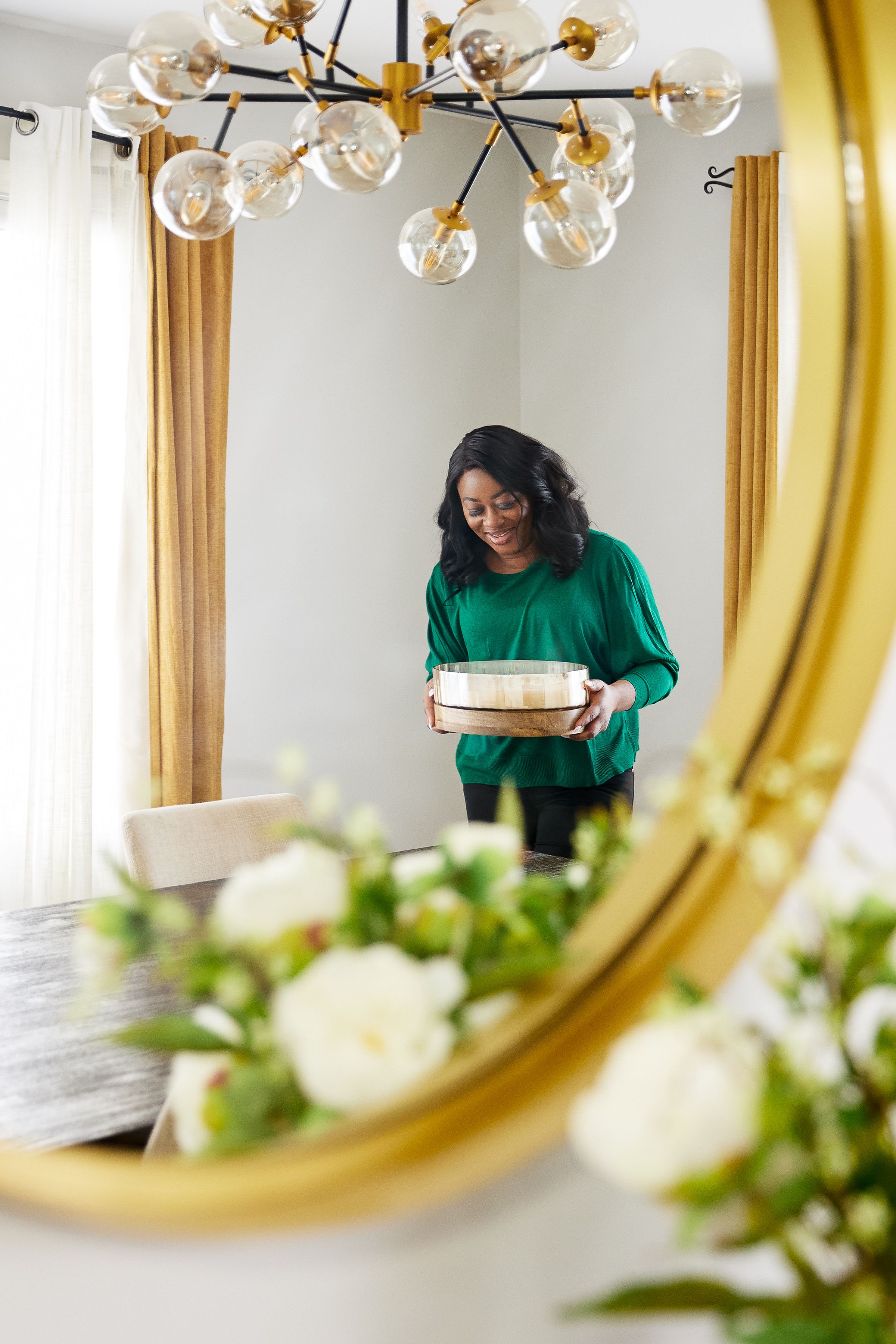 Woman in emerald green sweater arranges books on shelf near decorative gold mirror and crystal chandelier.