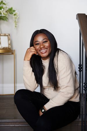 A person in a beige sweater and black pants poses casually on dark wooden stairs in a bright interior setting.
