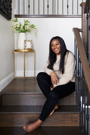 Woman in cream sweater and black pants sits casually on modern wooden staircase with black railing and white walls.