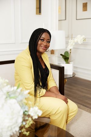 Professional wearing a bright yellow pantsuit sits on a chair in an elegant room with white flowers and decor.