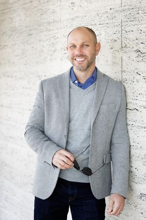 Professional headshot of a person wearing a light gray blazer and blue sweater against a textured wall background.
