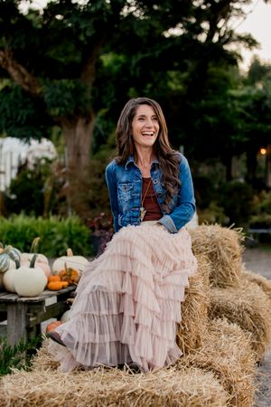 A woman in a denim jacket and pink tulle skirt sits on hay bales surrounded by autumn pumpkins and decor.