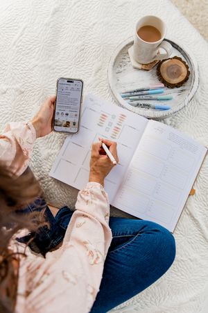 A person in blue jeans and pink shirt writes in a planner while having coffee on a white textured surface.