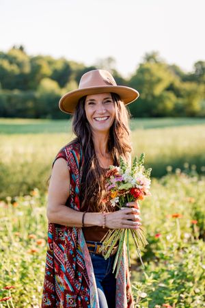 A person wearing a brown hat and floral dress picks wildflowers in a sunny meadow during golden hour.