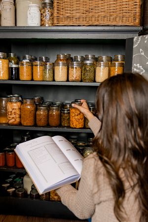Someone consults a recipe book while organizing preserved foods in mason jars on dark wooden shelves.