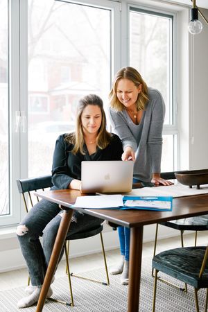 Two people collaborating at a modern office desk with laptops and paperwork in a bright windowed space.