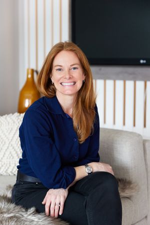 Professional portrait of someone in a navy blue top sitting comfortably on a gray couch in a bright modern room.