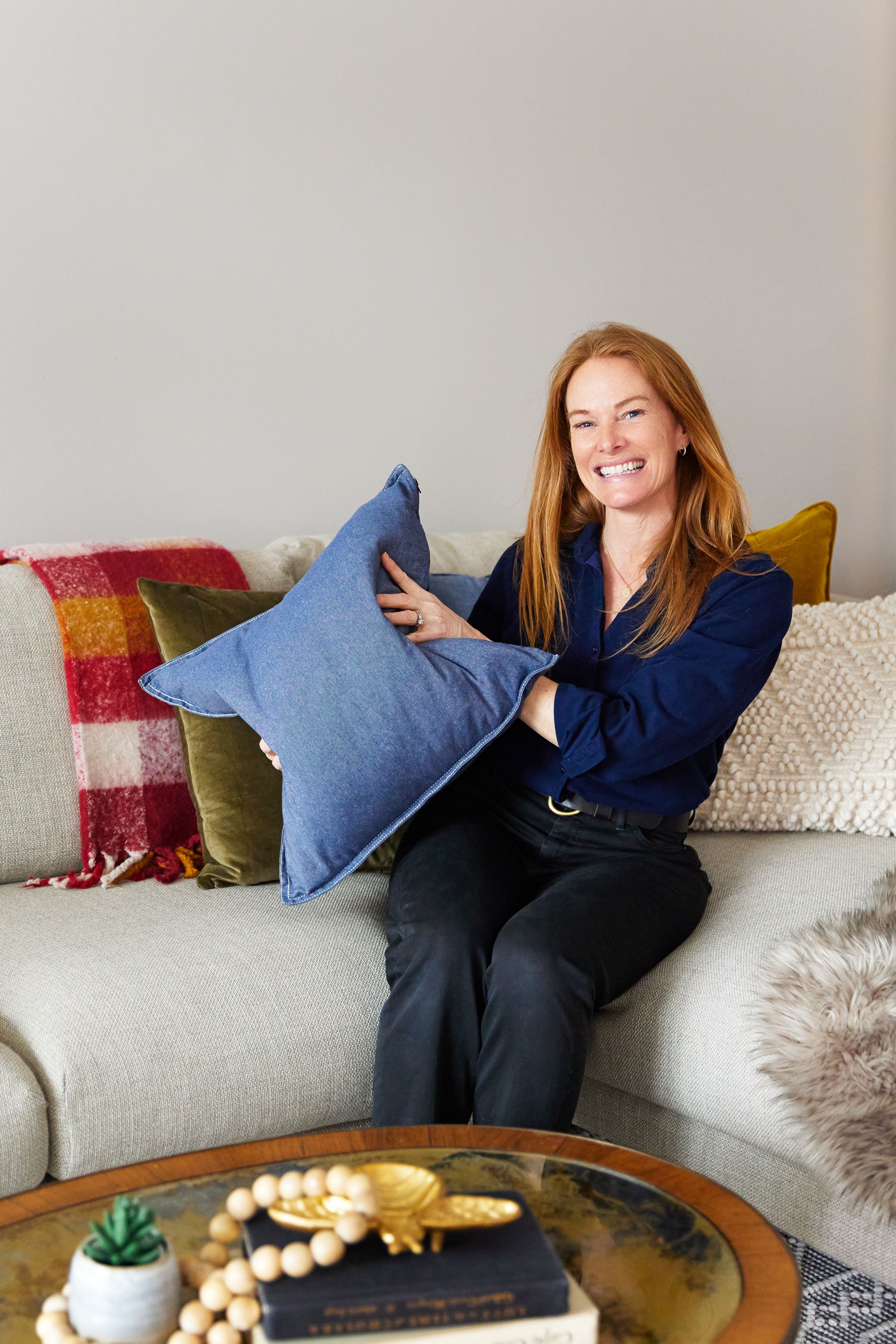 Relaxed woman sitting on gray couch with colorful throw pillows and red plaid blanket in cozy living room setting.