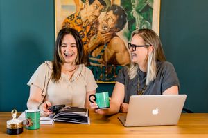 Two coworkers share a laugh while working together at a wooden table with laptops and coffee mugs.