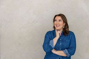 Person in denim shirt poses thoughtfully against light gray wall while looking up and smiling.