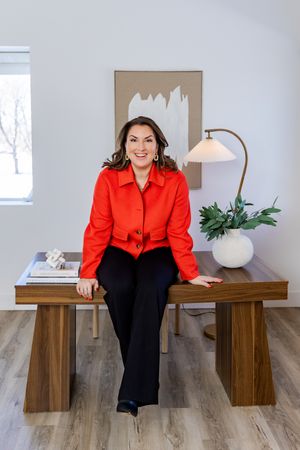 A person in a vibrant red blouse and black pants sits on a wooden bench in a minimalist interior setting.