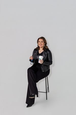 A person in a black outfit sits casually on a white stool against a light background, holding a white mug.