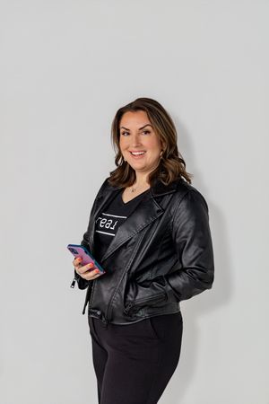 Professional wearing a black leather jacket and dark pants holds a phone while posing against a light gray background.