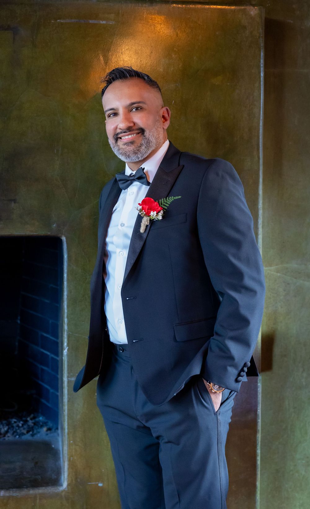 A groom in a classic black tuxedo and bowtie with a red boutonniere poses against a textured gold wall.