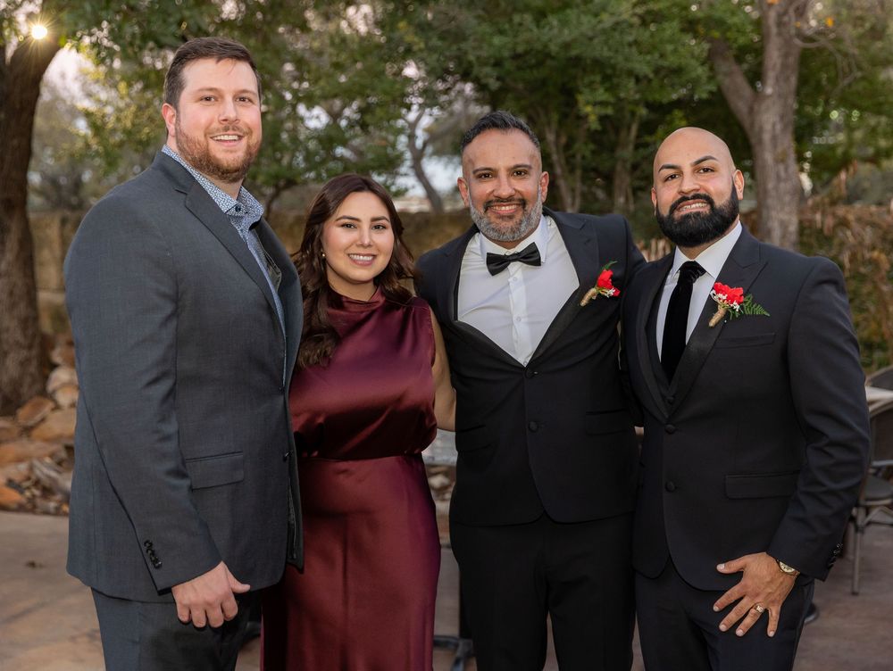 A group in formal attire poses together at a wedding, with men in dark suits and a woman in burgundy dress.