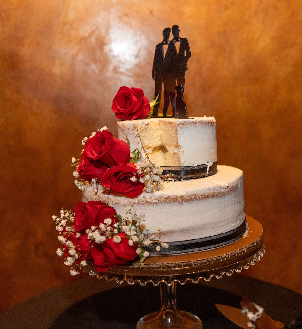 Wedding cake decorated with red roses and baby's breath on a gold stand against a warm copper-colored background.