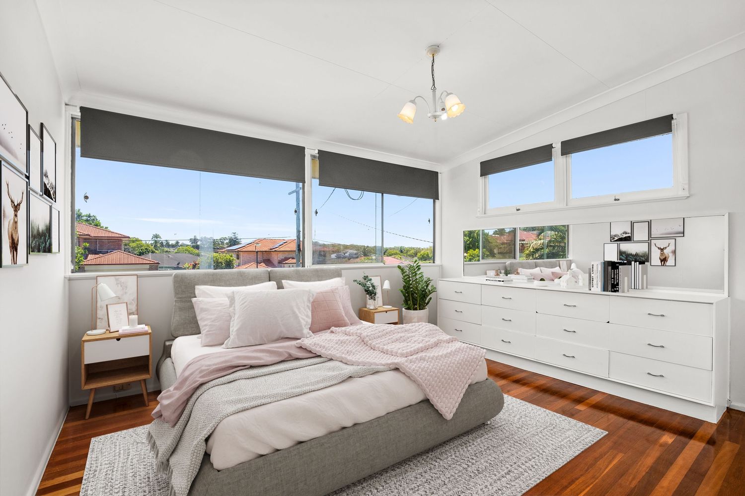 Bright master bedroom with hardwood floors, large windows, pink bedding, white dresser, and panoramic suburban views.