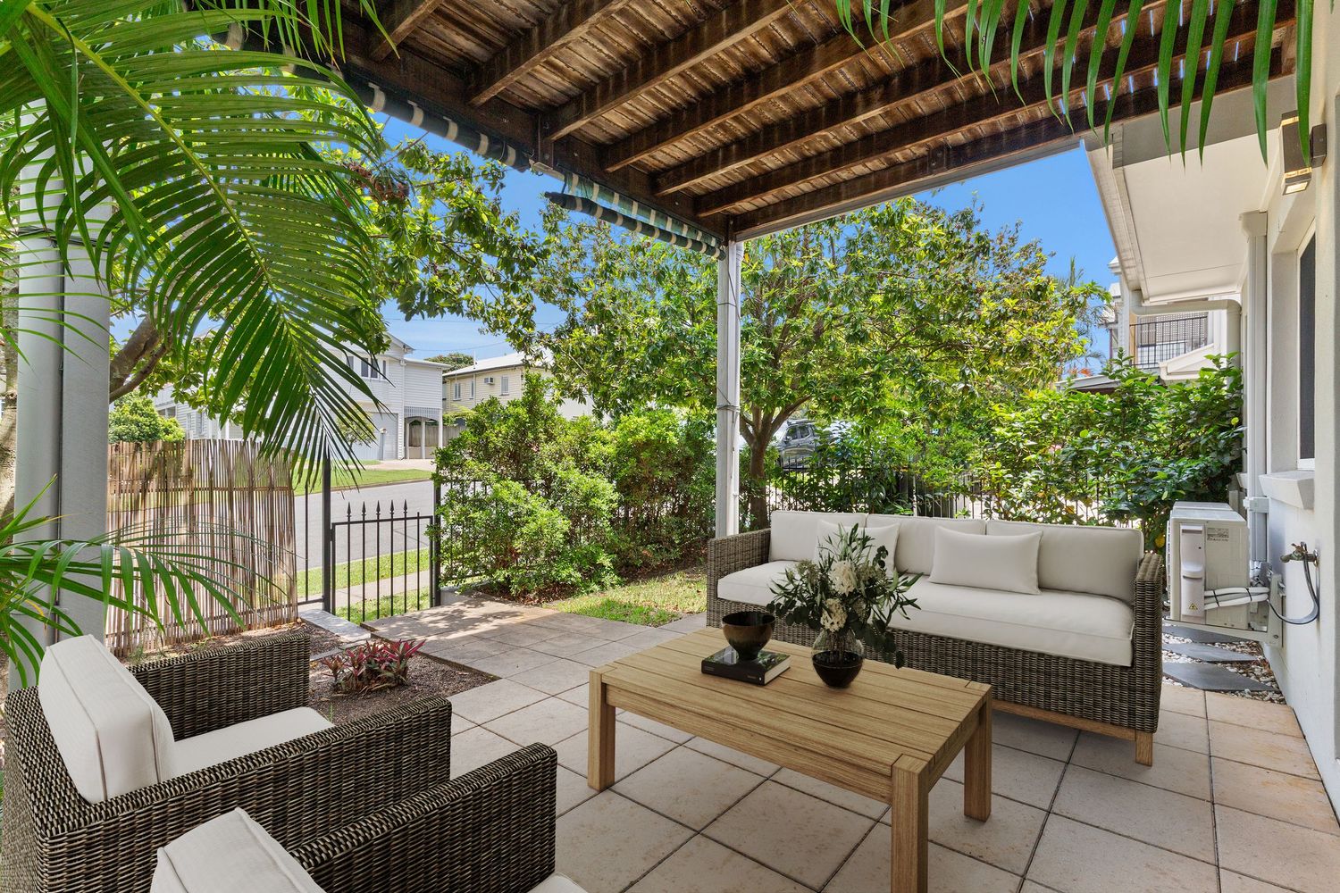 Elegant outdoor patio with wicker furniture, white cushions, timber coffee table, and lush garden views under a timber pergola.
