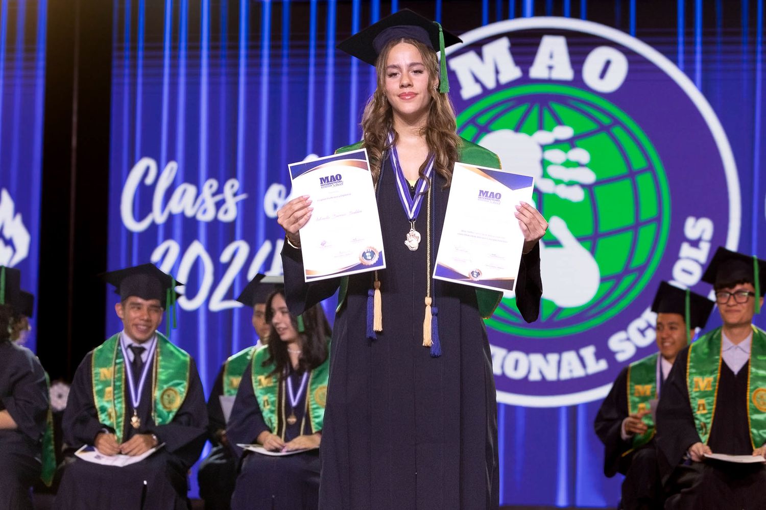 Award recipients stand on stage with medals and certificates at a formal ceremony with blue lighting.