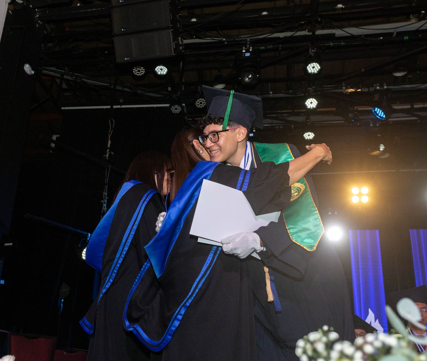 A stage performance with blue lighting and a figure in a graduation cap and gown speaking at a podium.