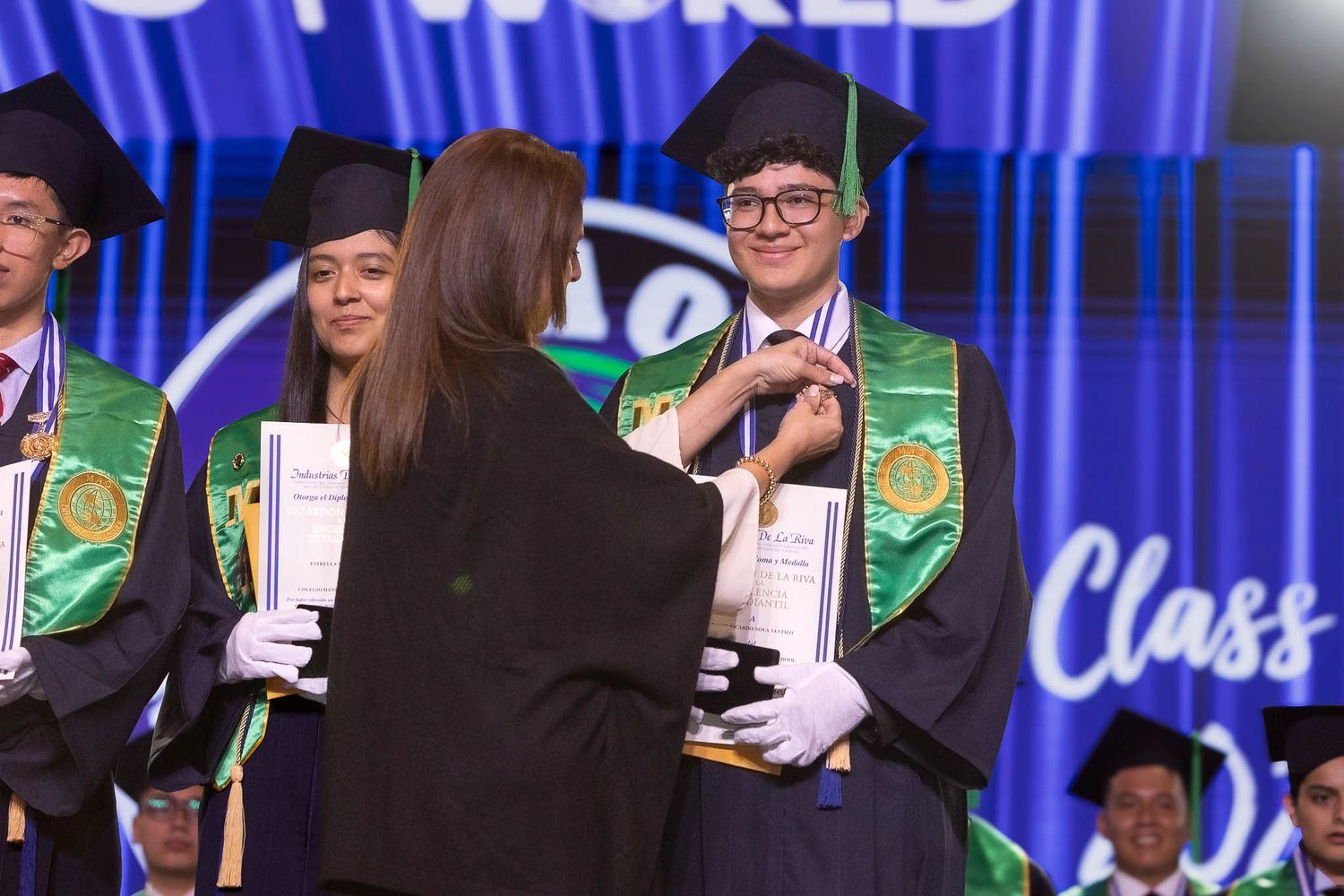 Students in green stoles receiving diplomas during graduation ceremony with blue backdrop.