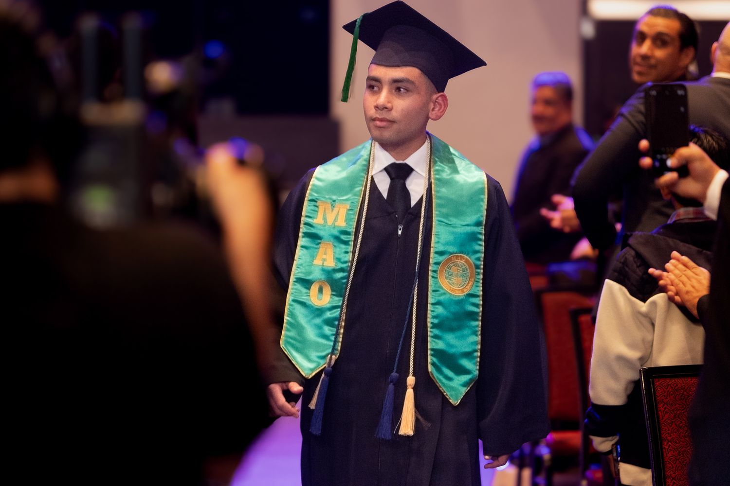 Graduate wearing navy cap and gown with decorative teal and sunflower graduation stole at commencement ceremony.